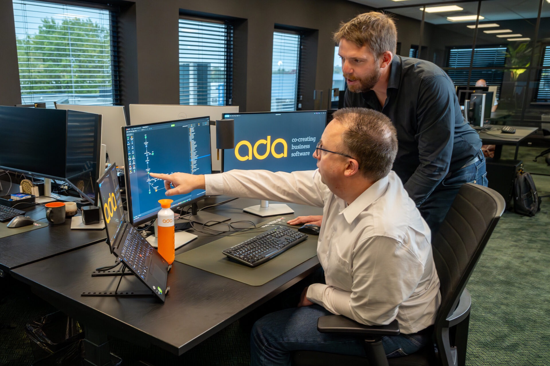 Twee ADA specialisten bekijken samen een workflow op meerdere schermen tijdens de ontwikkeling van bedrijfssoftware.